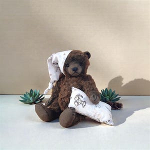 Puede incluir: Un oso de peluche marrón con un gorro de dormir blanco con un estampado floral, sosteniendo una pequeña almohada con una ilustración de tigre. El oso está sentado junto a una planta suculenta. El pelaje del oso es de color marrón oscuro.