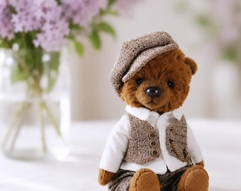 May include: A brown plush teddy bear wearing a white collared shirt, a brown herringbone patterned waistcoat with three buttons, matching trousers, and a matching flat cap. A small gold-toned chain is attached to the front of the waistcoat.