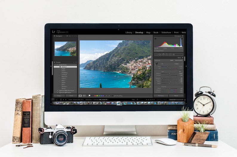 May include: A computer screen displaying the Adobe Lightroom CC software with a photo of a coastal town in Italy. The screen shows the Develop module with various editing tools and a histogram. A camera, a keyboard, and a clock are on the desk in front of the computer.