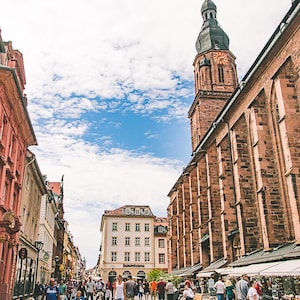 May include: A street scene in a European city with a tall church tower in the background. The street is lined with buildings and there are people walking around. The sky is blue with white clouds.