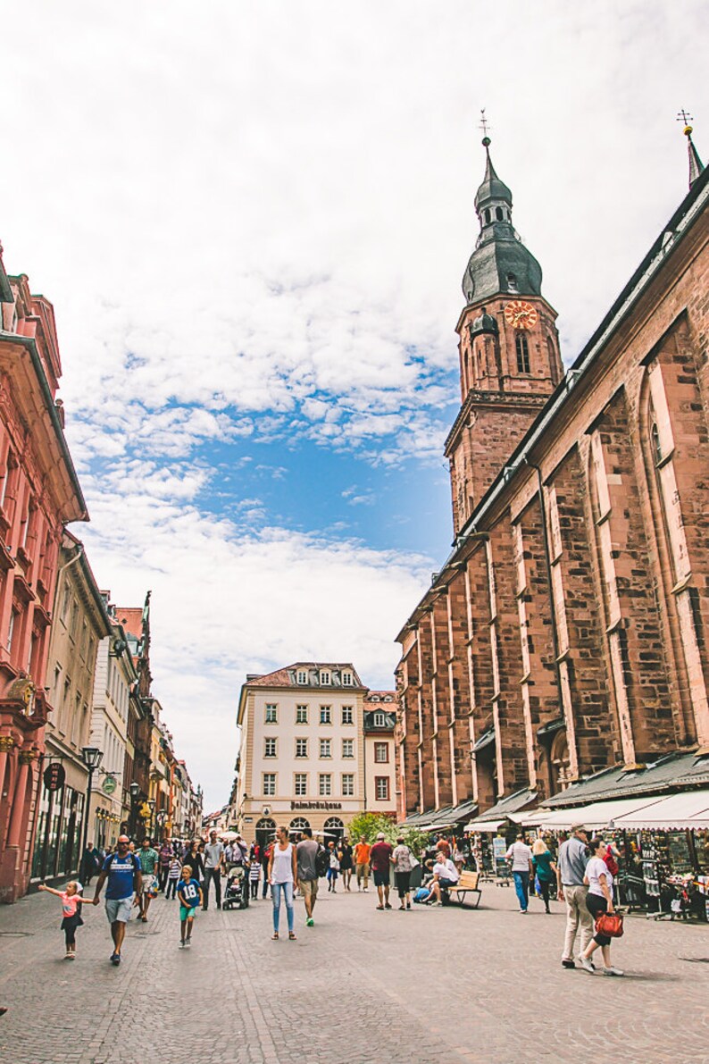 May include: A street scene in a European city with a tall church tower in the background. The street is lined with buildings and there are people walking around. The sky is blue with white clouds.