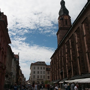 May include: A street scene in a European city with a tall church tower in the background. People are walking along the cobblestone street, and there are shops and buildings on either side.