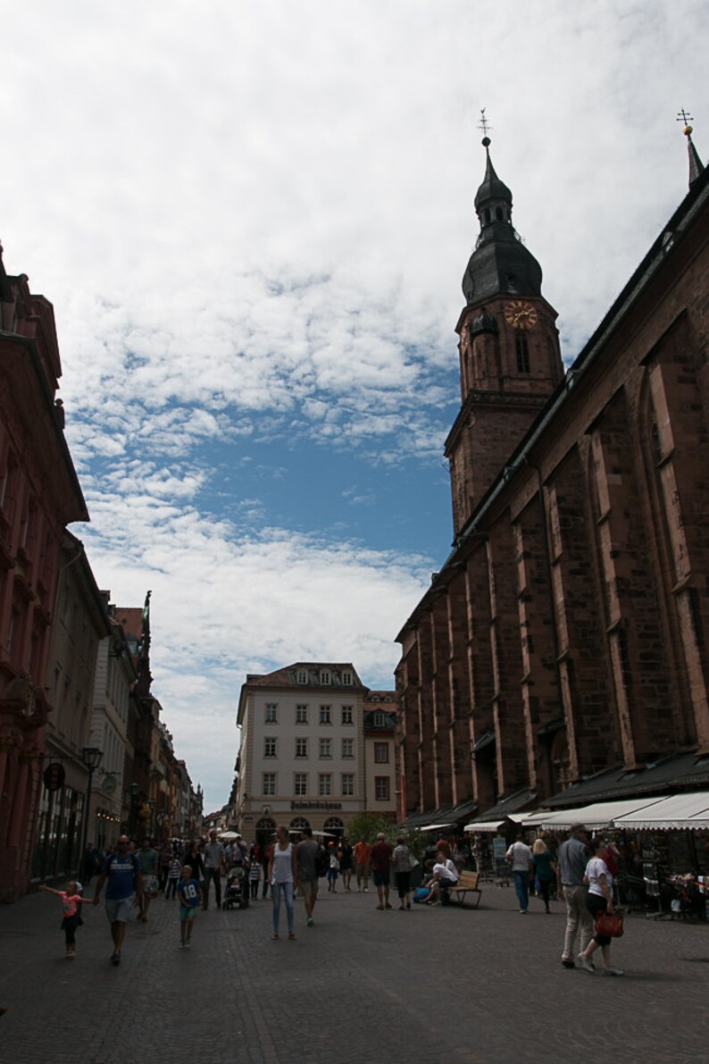 May include: A street scene in a European city with a tall church tower in the background. People are walking along the cobblestone street, and there are shops and buildings on either side.