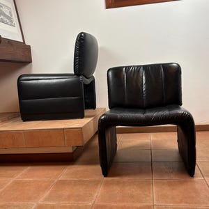 May include: Two black leather armchairs with a distinctive, curved design. One chair has a separate backrest, while the other is a single piece. The chairs are placed on a tiled floor, showcasing a modern aesthetic.