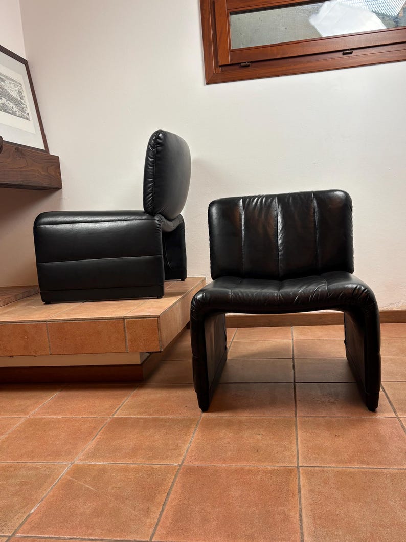 May include: Two black leather armchairs with a distinctive, curved design. One chair has a separate backrest, while the other is a single piece. The chairs are placed on a tiled floor, showcasing a modern aesthetic.