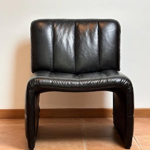 May include: A black leather chair with a low back and a curved seat. The chair has a rectangular back with vertical stitching and a rounded seat. The chair is made of black leather and has a modern design.