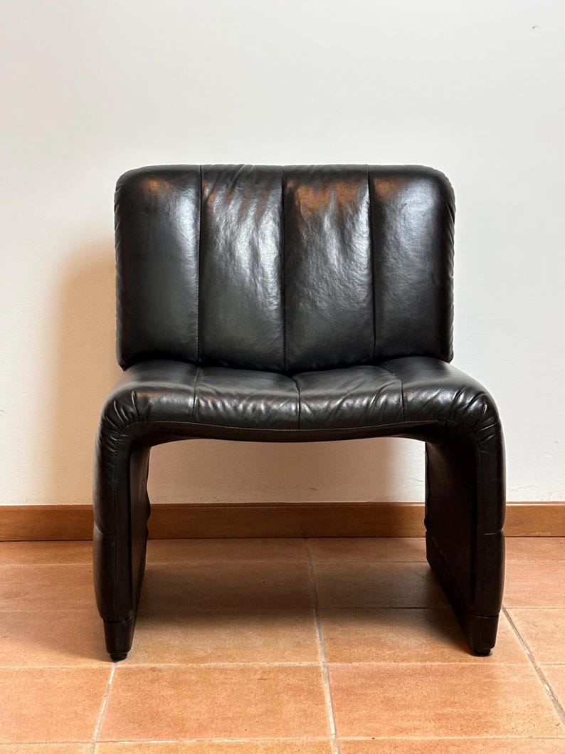 May include: A black leather chair with a low back and a curved seat. The chair has a rectangular back with vertical stitching and a rounded seat. The chair is made of black leather and has a modern design.