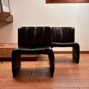 May include: Two black leather armless chairs with a curved base and a segmented backrest. The chairs are positioned on a terracotta tile floor, with a wooden window frame visible in the background.