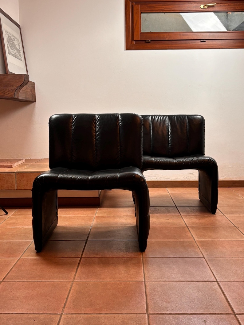 May include: Two black leather armless chairs with a curved base and a segmented backrest. The chairs are positioned on a terracotta tile floor, with a wooden window frame visible in the background.