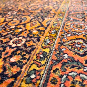 May include: Close-up of an orange and navy rug with a detailed floral pattern. The rug has a central design with a border, featuring yellow, green, and white accents. The rug's texture is visible, showcasing its woven construction.