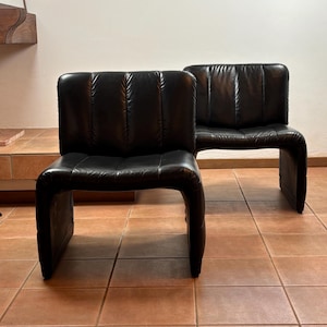 May include: Two black leather armless chairs. The chairs have a curved, continuous frame and a padded backrest with vertical stitching. The chairs are placed on a tiled floor.