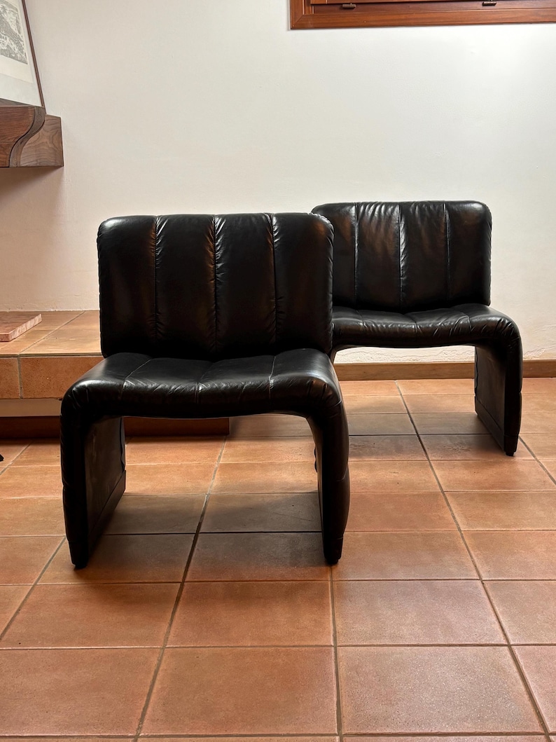 May include: Two black leather armless chairs. The chairs have a curved, continuous frame and a padded backrest with vertical stitching. The chairs are placed on a tiled floor.
