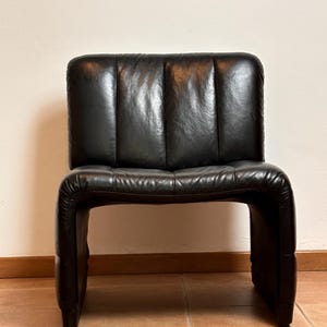 May include: A black leather chair with a distinctive, curved design. The chair has a rectangular backrest with vertical stitching and a seat that curves into the legs. The chair is made of a dark, glossy leather.