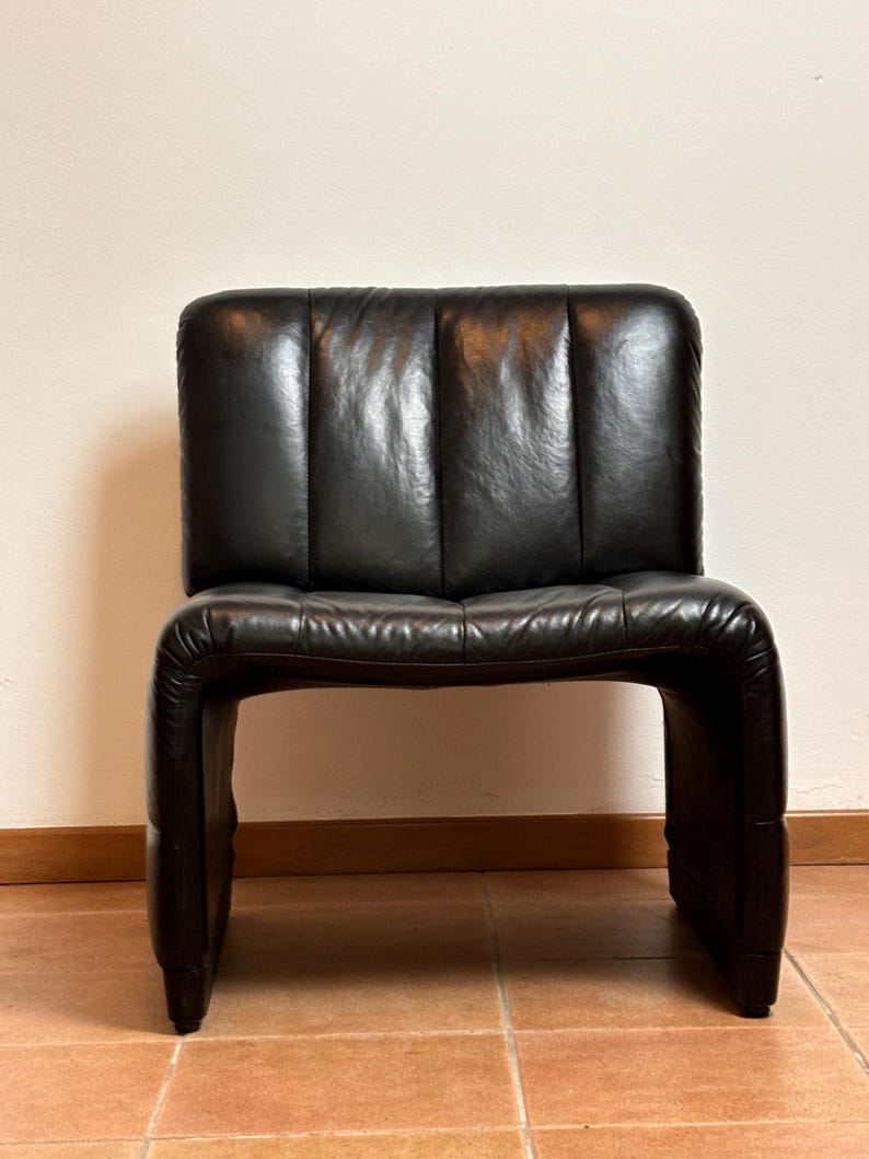 May include: A black leather chair with a distinctive, curved design. The chair has a rectangular backrest with vertical stitching and a seat that curves into the legs. The chair is made of a dark, glossy leather.