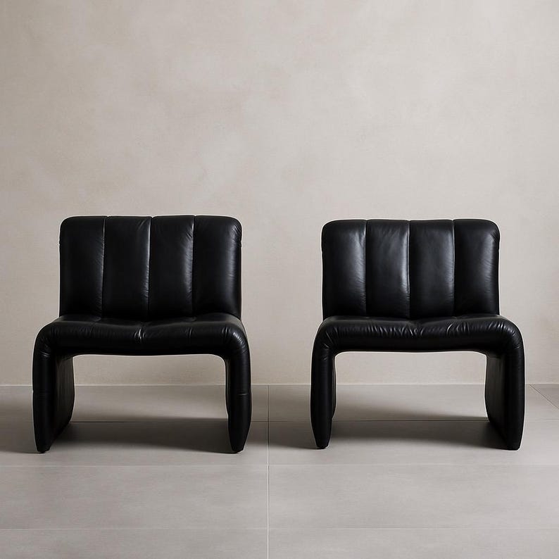 May include: Two black leather chairs with a contemporary design. Each chair features a padded back with vertical stitching and a curved, continuous frame. The chairs are set against a neutral backdrop.