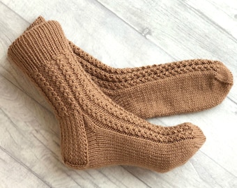 May include: A pair of brown knitted socks with a cable knit pattern. The socks are lying on a light wood surface.