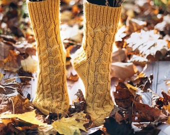 May include: A pair of yellow knitted socks with a cable knit pattern. The socks are worn by a person standing in a pile of autumn leaves.