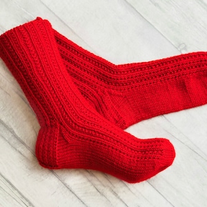 Puede incluir: Un par de calcetines de punto rojo con un patrón de canalé. Los calcetines están doblados y colocados sobre una superficie de madera blanca.