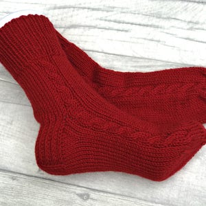 May include: A pair of red knitted socks with a cable knit pattern. The socks are folded over and the top of the socks are white.