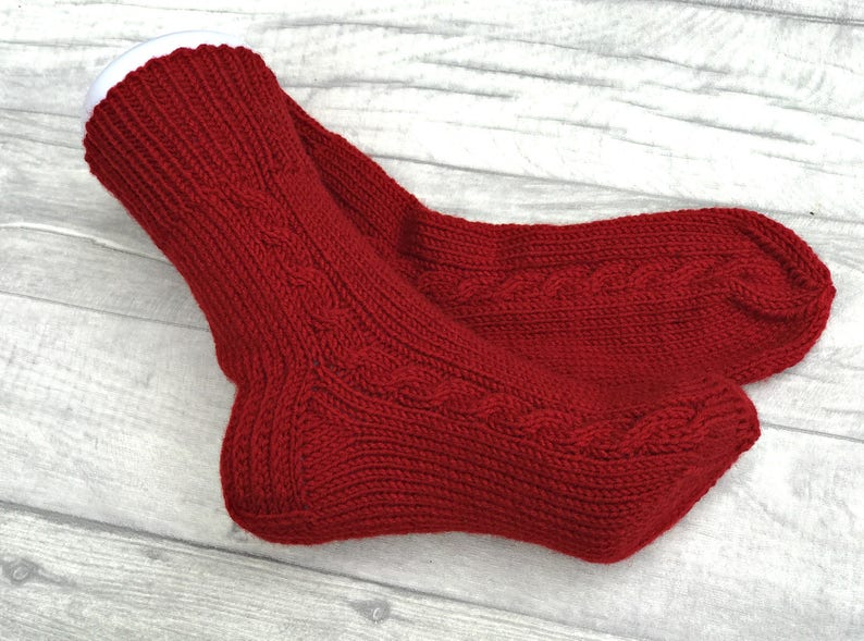 May include: A pair of red knitted socks with a cable knit pattern. The socks are folded over and the top of the socks are white.