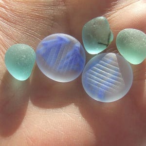 Op de afbeelding: Een verzameling van vijf gladde, matglazen stukjes zeeglas. Twee zijn rond met reliëflijnen in blauw en wit, terwijl de andere drie onregelmatige, doorschijnende vormen hebben in tinten van lichtblauw en bleek zeegroen.