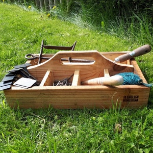 May include: A wooden toolbox containing gardening tools such as a trowel and twine, resting on grass. The toolbox has a handle and compartments. The text "AN ENGLISH HOMESTEAD" is printed on the side. Various metal tools are also visible.