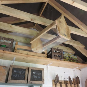 May include: A wooden bird feeder with a wire mesh bottom hangs from the ceiling of a wooden shed. The feeder is made of unfinished wood and has a simple design. The shed has a white painted wall and a wooden shelf with various items on it, including a wooden crate, a box labeled "Catalogue of Garden Seeds", and a chalkboard sign that says "Hosepipe Fittings".