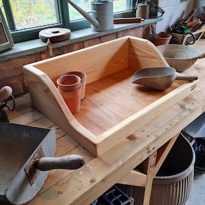 May include: A wooden tray with a scoop and three terracotta pots. The tray is sitting on a wooden workbench with a metal bucket and a black hose.