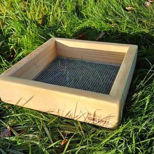 May include: A wooden sifter box with a fine mesh screen base, set on green grass. The box is rectangular with a light wood finish and a logo on the side. The sifter is designed for gardening or crafts.