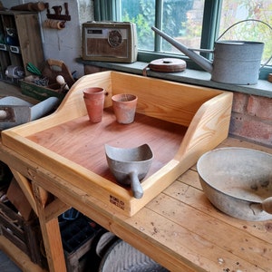 May include: A wooden potting bench with a built-in tray, two terracotta pots, a metal scoop, and a metal bowl with a wooden handle. The bench is made of unfinished wood and has a label that reads "Made in England".
