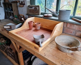 May include: A wooden potting bench with a built-in tray, two terracotta pots, a metal scoop, and a metal bowl with a wooden handle. The bench is made of unfinished wood and has a label that reads "Made in England".