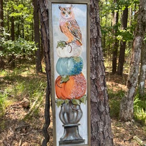 May include: A rectangular framed painting featuring a vertical stack of three pumpkins in white, light blue, and orange, topped with a brown owl. The pumpkins rest on a dark gray pedestal base, with green leaves tucked between each layer.