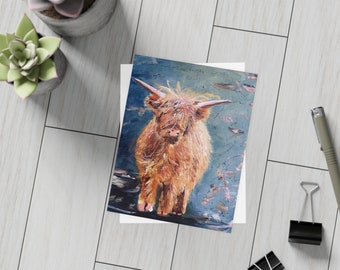 May include: A greeting card featuring a painting of a Highland cow. The artwork depicts the cow with long, shaggy, reddish-brown fur and long horns against a blue background. The card is on a white surface with succulents and office supplies.