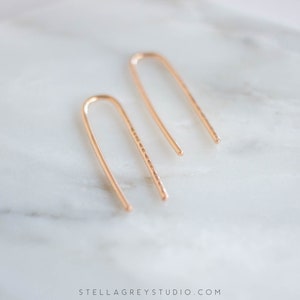 May include: A pair of gold-toned minimalist earrings with a hammered texture. The earrings are shaped like a thin, curved line.