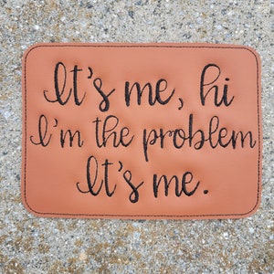 May include: A brown leather coaster with black embroidered text that reads "It's me, hi I'm the problem It's me."