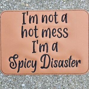 May include: A brown leather patch with black embroidered text that reads "I'm not a hot mess I'm a Spicy Disaster".