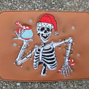 Peut inclure: Patch brodé représentant un squelette portant un bonnet de Père Noël et tenant une tasse de chocolat chaud avec une canne en sucre. Le squelette est entouré de lumières de Noël et de flocons de neige.