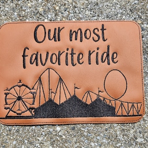 May include: Brown leather patch with black embroidery. The embroidery depicts a roller coaster, Ferris wheel, and carnival tents. The text "Our most favorite ride" is embroidered above the carnival scene.