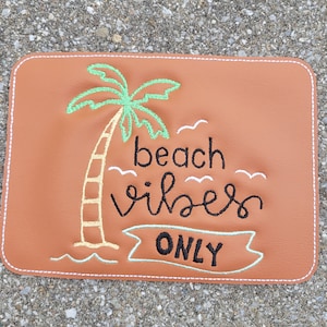 May include: A brown leather patch with white stitching, featuring an embroidered green palm tree and the text "beach vibes only".