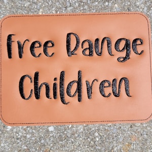 May include: A rectangular, tan-coloured patch with the words "free Range Children" embroidered in black, cursive lettering. The patch has a black stitched border.
