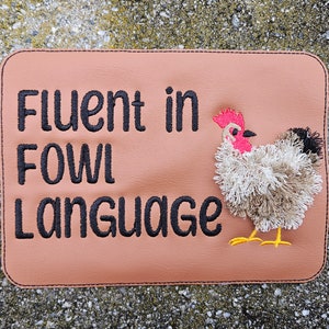 May include: A rectangular, tan-colored patch with the phrase "Fluent in Fowl Language" embroidered in black. A decorative chicken with a red comb, black and white feathers, and yellow feet is on the right side.