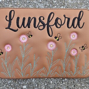 May include: A brown rectangular patch with the name "Lunsford" embroidered in black script. The patch features embroidered pink flowers with yellow centers and small black and yellow bees. The flowers have green stems.