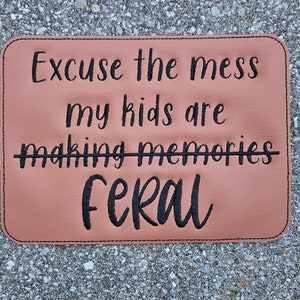 May include: A brown leather doormat with black embroidered text that reads "Excuse the mess my kids are making memories feral".