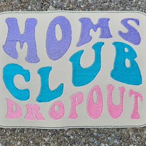 May include: A beige fabric patch with the text "Moms Club Dropout" in colorful embroidered letters. The letters are in shades of pink, blue, and purple.