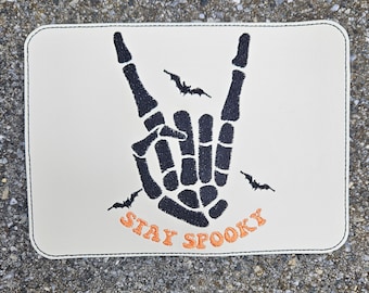 May include: A white patch with black embroidery of a skeleton hand making the rock and roll sign. There are bats flying around the hand. The text "STAY SPOOKY" is embroidered in orange below the hand.