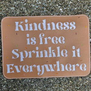 May include: A tan leather pouch with a stitched border and the text "Kindness is free Sprinkle it Everywhere" embroidered in white.