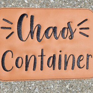 May include: A brown leather coaster with black embroidery that reads "Chaos Container".