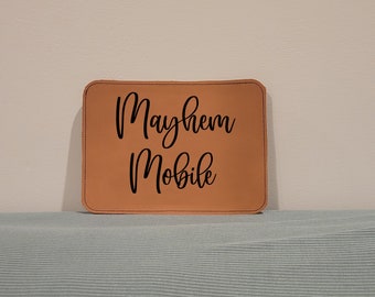 Op de afbeelding: Een bruin leren patch met de tekst "Mayhem Mobile" in zwarte cursieve letters.