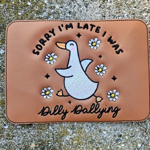 May include: A brown leather patch with white embroidery of a goose with the text "Sorry I'm late I was dilly dallying" and white flowers.