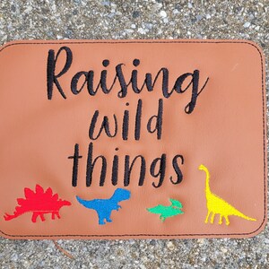 Peut inclure: Sous-verre en cuir marron avec le texte "Raising wild things" brodé en noir. Il y a quatre silhouettes de dinosaures colorées brodées sur le sous-verre.